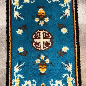 May include: Rectangular turquoise rug with a detailed pattern. The design includes white, yellow, and brown floral and geometric motifs. The rug has a black border and fringe.