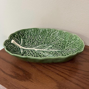 May include: An oval ceramic serving dish shaped like a large green cabbage leaf. The bowl features a glossy finish with a textured surface that mimics the veins and ruffled edges of a real leaf, with a prominent white stem running through the center.