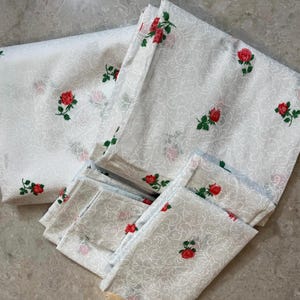 May include: White bedding set with a delicate floral pattern of red roses and green leaves. The set includes a folded sheet, a flat sheet, and several pillowcases. The fabric has a subtle, swirling design.