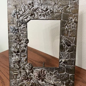 May include: A rectangular picture frame with a dark bronze finish featuring a raised relief design of climbing roses, leaves, and vines on the left side, and a textured stone wall pattern with small insect details on the right side.