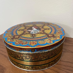 May include: An oval tin box with a decorative design. The lid features a central image of a woman in a portrait, surrounded by gold, blue, and red patterns. The sides of the tin are adorned with gold and green bands.