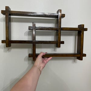 May include: A dark brown wooden wall shelf featuring an interlocking rectangular frame design. The structure consists of multiple horizontal and vertical wooden slats joined together to create several open storage compartments with a glossy finish.