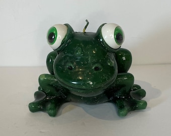 May include: A green frog candle with white eyes. The candle is sitting on a white surface.