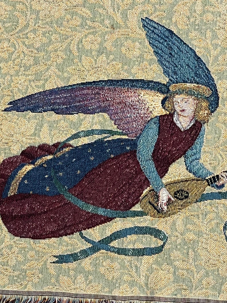 Puede incluir: Tapiz que representa a un &aacute;ngel tocando un la&uacute;d. El &aacute;ngel tiene alas azules y moradas, un vestido granate y una blusa azul. El fondo es un estampado floral en tonos verdes y beige. El tapiz tiene un borde con flecos.