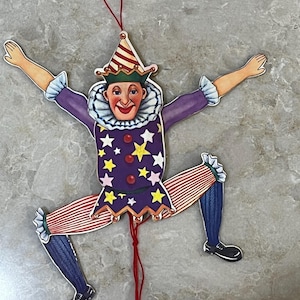 May include: A colourful clown ornament with outstretched arms and legs. The clown has a purple jacket with yellow stars, red and white striped legs, and a red string for hanging. The clown's face has a cheerful expression.
