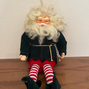 May include: A Christmas elf doll with a white beard and hair, wearing a dark green jacket, red and white striped pants, and green shoes. The doll has a gold-colored belt and is sitting on a wooden surface.