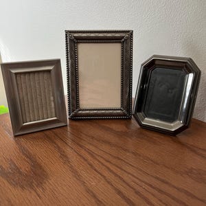 May include: Three decorative picture frames in varying shapes and sizes. The frames are in shades of brown and bronze, with one rectangular frame featuring a textured border. The frames are displayed on a wooden surface.