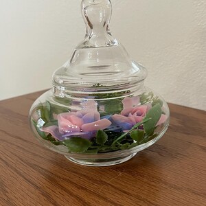May include: A clear glass jar with a lid, containing pink and blue roses and green leaves. The jar sits on a wooden surface. The roses are a focal point, adding a touch of color and elegance to the decorative piece.
