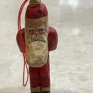 May include: A hand-carved wooden Santa Claus Christmas ornament. The ornament is painted red with a white beard and a red hat. A red string is attached for hanging. The Santa has a stern expression.