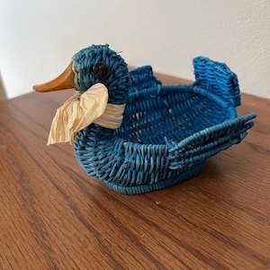 May include: A blue woven duck-shaped basket with a wooden beak and a cream-colored ribbon tied around its neck. The basket has a round body and wing-shaped sides. The duck's eye is a dark, round button.