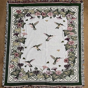 May include: A woven throw blanket featuring a light blue background with a floral and hummingbird design. The border is green with a geometric pattern and fringe. The blanket has a repeating pattern of hummingbirds and flowers.