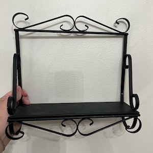 May include: A black metal wall shelf featuring a flat rectangular base and a decorative frame with scrolled wire accents at the top and bottom. The structure includes two vertical side supports with curved detailing connecting the shelf to the top.