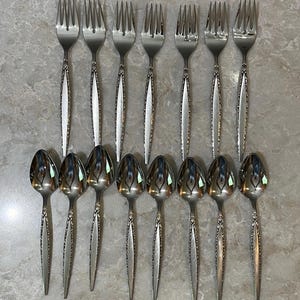 May include: A set of seven stainless steel forks and eight stainless steel spoons arranged in two rows. Each piece of flatware features a polished finish on the head and a textured, tapered handle with a decorative border design.