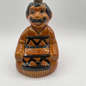 May include: A brown ceramic figurine of a man with a mustache and a patterned shirt. The figurine has a cork stopper on top of its head.