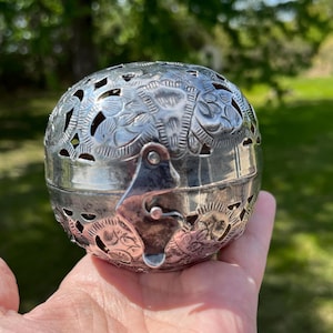 May include: A silver-colored, spherical container with intricate floral and geometric cut-out designs. The container has a hinged lid with a clasp closure. The surface is detailed with embossed patterns. The object is held in a hand.