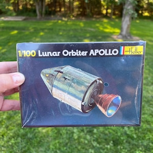 May include: A model kit box for a 1/100 scale Lunar Orbiter Apollo spacecraft. The box features a detailed illustration of the spacecraft against a dark blue background. The Heller brand logo is visible.