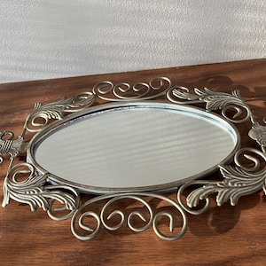 May include: An oval vanity mirror with an ornate, antique-style metal frame. The frame features decorative scrollwork and leaf-like accents. The mirror is set on a wooden surface, reflecting the light.