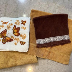 May include: A selection of vintage towels in brown and gold hues. One white towel is embroidered with butterflies and floral patterns. A dark brown towel with a lace trim is also present. The towels are displayed on a light-coloured surface.