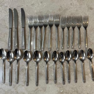 May include: A collection of vintage silverware, including knives, forks, and spoons, arranged on a light-colored surface. The silverware has a silver finish with decorative detailing on the handles. The set includes various sizes of spoons and forks.