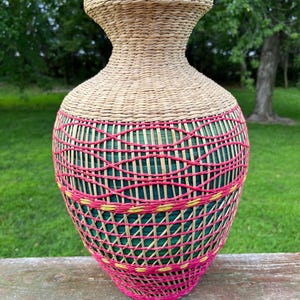 May include: A tall, bulbous vase crafted from woven natural tan fibers. The upper section features a tight weave, while the lower body is encased in a decorative lattice of bright pink and yellow fibers over a solid dark green inner layer.