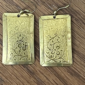 May include: A pair of rectangular gold-toned metal earrings featuring an intricate embossed floral and scrollwork pattern. Each earring is suspended from a simple metal hook wire, showcasing a polished finish with recessed details in the design.
