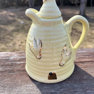 May include: A pale yellow ceramic honey pot with a lid, handle, and spout. The pot is shaped like a beehive with gold-colored bee accents. The beehive has a small opening at the bottom.