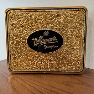 May include: A rectangular gold-colored Whitman's Sampler candy tin. The tin has an embossed floral design and a black oval label with the brand name in white script. The tin is sitting on a wooden surface.