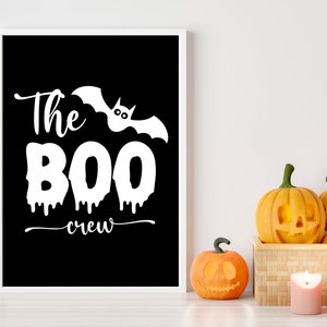 May include: A black and white printable sign with the text "The BOO crew" and a white bat with eyes. The sign is framed in a white frame and is sitting on a white surface with three carved pumpkins.