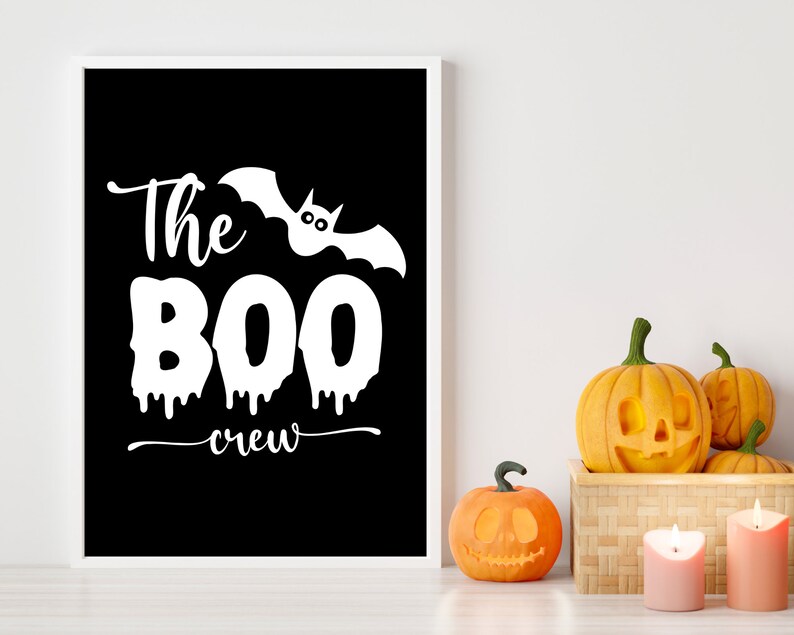 May include: A black and white printable sign with the text "The BOO crew" and a white bat with eyes. The sign is framed in a white frame and is sitting on a white surface with three carved pumpkins.