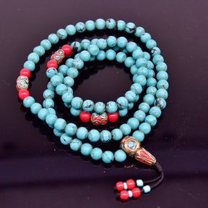 May include: A turquoise and red beaded mala necklace with a gold-toned tassel. The beads are round and have a slightly textured surface. The mala is a traditional Buddhist prayer bead necklace.