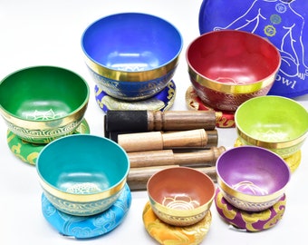 May include: Set of seven singing bowls with colorful exteriors and gold trim. Each bowl is on a matching cushion and comes with a wooden striker. The bowls are arranged in a circle with a blue carrying case in the background.