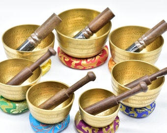 Op de afbeelding: Een verzameling van zeven gouden zangschalen met houten stokken. Elke kom rust op een kleurrijk kussen. De kommen hebben een gehamerde textuur en zijn gerangschikt op een witte achtergrond, wat duidt op meditatie en klankgenezing.