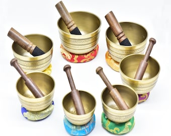 Op de afbeelding: Zes gouden zangkommen met houten slagers, elke kom rust op een kleurrijk, gepatroneerd kussen. De kommen hebben ingewikkelde ontwerpen en zijn in een halve cirkel op een witte achtergrond gerangschikt.