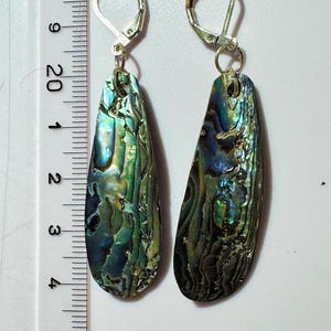 May include: A pair of iridescent abalone shell earrings. The earrings have a teardrop shape, with silver-coloured leverback closures. The shells show green, blue, and gold colours, with a textured surface. Each earring is approximately 5 cm long.
