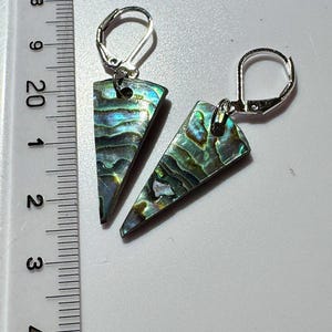 May include: A pair of iridescent abalone shell earrings with silver-colored lever-back closures. The earrings are triangular, with a shimmering mix of green, blue, and purple hues. The ruler next to the earrings shows a measurement of approximately 1 inch.