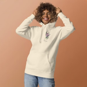 May include: Cream-coloured hoodie with a front pocket and drawstring hood. The hoodie features a floral design with the word "Mom" printed below the flowers. The person is wearing blue jeans.