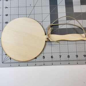 Puede incluir: Un disco de madera en blanco en forma de círculo con un mango adjunto. El disco tiene un diámetro de aproximadamente 10 cm. El mango es de madera y tiene una longitud aproximada de 15 cm.