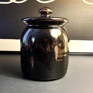 May include: A black ceramic jar with a lid. The jar has a glossy finish and a rounded shape. The lid is topped with a small, round knob. The jar is sitting on a dark surface.
