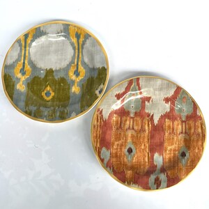May include: Two decorative ceramic plates with a gold rim. One plate features a pattern in shades of grey, yellow, and green. The other plate has a pattern in shades of red, orange, and grey. Both plates have a unique ikat design.