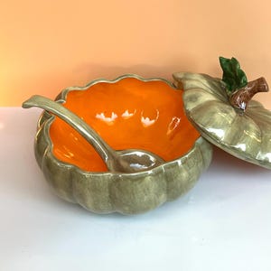 May include: A ceramic pumpkin-shaped tureen with a glossy sage green exterior and a vibrant orange interior. The set includes a matching lid with a brown stem and green leaf detail, along with a curved ceramic ladle resting inside the bowl.