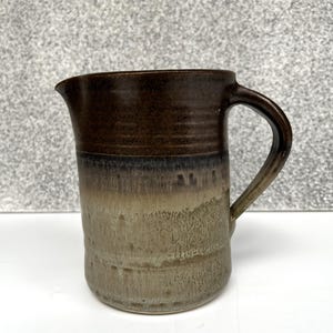 May include: A handmade ceramic jug with a dark brown top and handle, fading to a lighter, textured tan base. The jug has a spout for easy pouring and a curved handle. The stoneware jug is suitable for serving drinks.