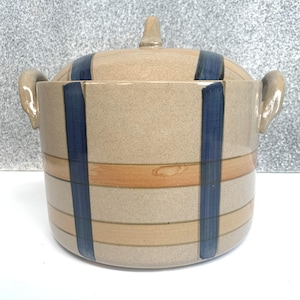 May include: A tan ceramic lidded jar with a cylindrical shape. The jar features horizontal bands of tan and orange, with vertical navy blue stripes. It has two side handles and a small knob on the lid.