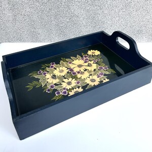 May include: A navy blue wooden serving tray with a floral design. The tray features a bouquet of yellow daisies and purple flowers on a dark green background. The tray has a rectangular shape with handles.
