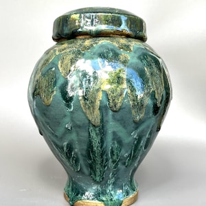May include: A teal and gold glazed ceramic jar with a lid. The jar has a rounded shape and a lid that fits securely. The glaze has a dripped effect, creating a textured appearance. The jar rests on a light-coloured base, adding to its aesthetic appeal.