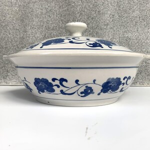 May include: White ceramic serving dish with a lid, decorated with blue floral and vine patterns. The dish has a rounded shape with a small handle on the lid and two small handles on the sides. A blue line accents the rim.