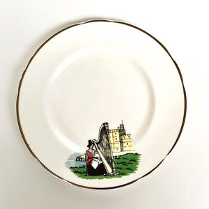 May include: A white ceramic saucer with a gold rim features a colourful illustration of a woman playing a harp in front of a castle. The scene is set against a green grassy hill with a blue sea in the background.