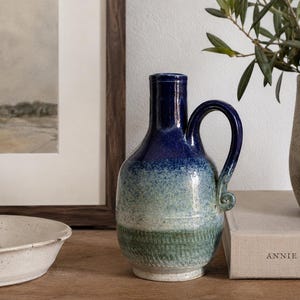May include: A ceramic pitcher featuring a gradient glaze that transitions from a deep navy blue at the neck to a lighter teal and sage green at the base. The vessel has a rounded body, a narrow neck, and a single curved handle with a curled end.