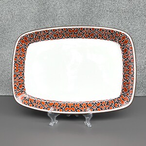 May include: Rectangular white serving platter with a decorative border. The border features a repeating pattern in shades of orange, black, and brown. The platter is displayed on a clear plastic stand.