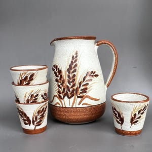 May include: A ceramic jug and four matching cups featuring a textured off-white finish with hand-painted brown wheat stalk motifs. The base of each piece is a solid brown band, and the jug includes a curved handle and a pouring spout.