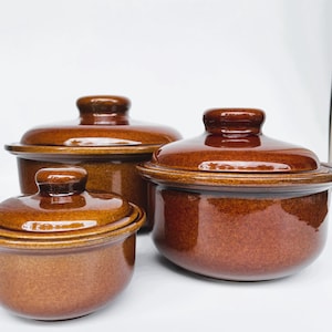 May include: Set of three brown ceramic casserole dishes with lids. The dishes are different sizes and have a glossy finish.